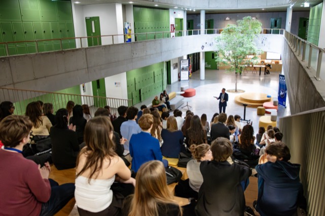 Participants seated on amphitheater steps listen to a speech in the school atrium.