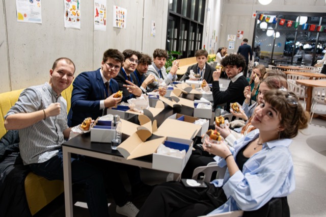 Delegates around a long dinner table wave and flash peace signs while eating burgers.