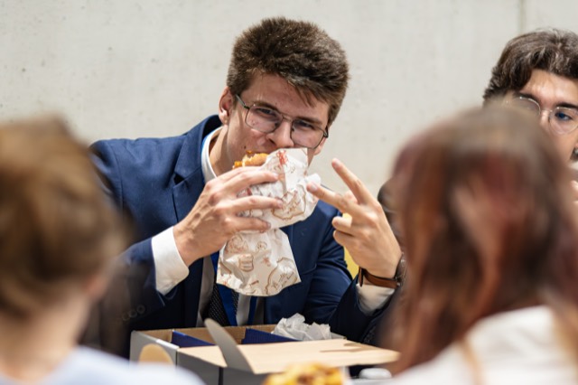 A delegate in glasses bites a burger while flashing a peace sign.