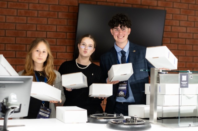 Three smiling students hold up stacks of white meal boxes behind the serving counter.