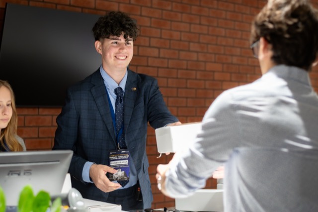 A cheerful organizer with a badge hands a white lunch box to a waiting delegate.