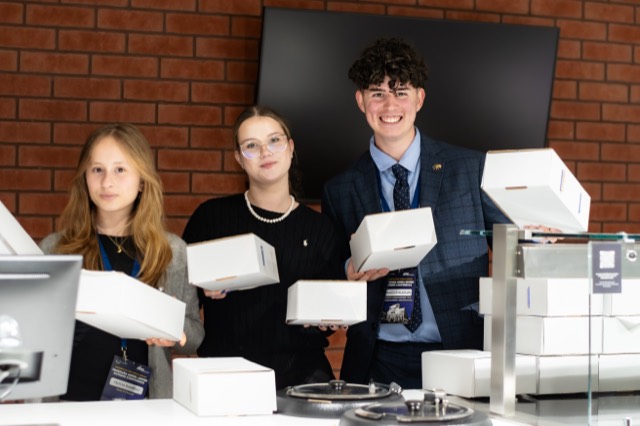 Three students grin while balancing several white meal boxes behind the counter.
