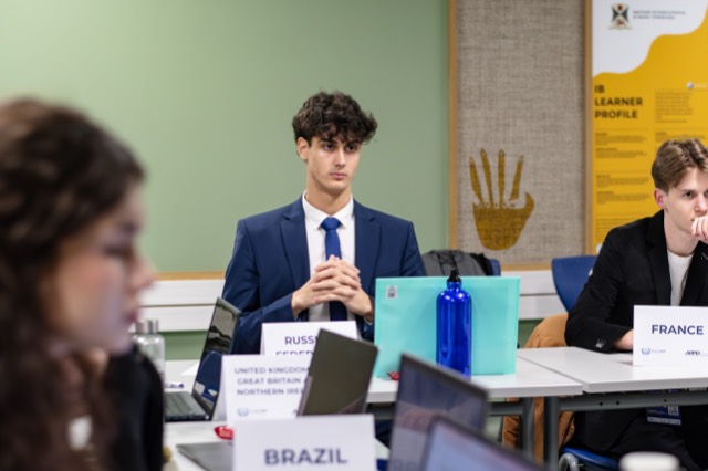 A delegate in a navy suit listens with hands clasped.