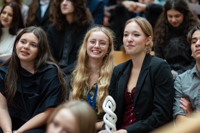 Three delegates smile in the audience, one holding a spiral award trophy.