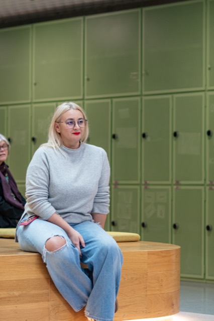 A woman in a grey sweater sits on a curved wooden bench in front of green lockers.