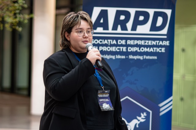 A conference official with an official badge speaks into a microphone in front of the ARPD banner.
