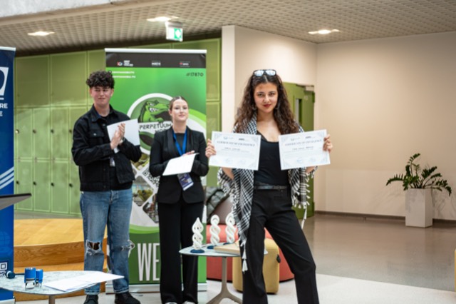 An award winner strikes a playful pose holding two certificates of excellence as others applaud.